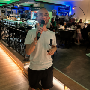 Il presentatore del Kanta Game canta al microfono davanti al bancone di 140 Grammi Sestu mentre i clienti seguono la serata musicale dai tavoli.